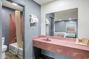 Vanity unit, mirror, wall shelf with towels, doorway to bathroom, shower tub with bath mat and curtain, toilet, laminate flooring