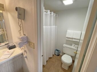 Vanity unit with towels, hairdryer, mirror, doorway leading to shower tub with shower curtain, toilet, hanging rail with towels, tiled flooring