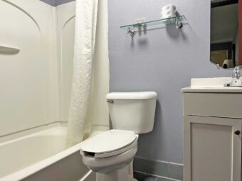 Showertub with shower curtain, toilet, shelf with bathroom amenities, vanity unit, mirror, tiled flooring