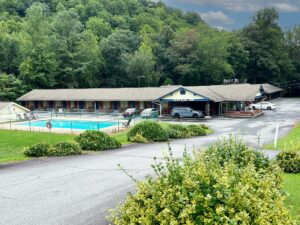 Outdoor pool and pool chairs surrounded by fencing, one story building with exterior guest room entrances, hotel entrance, parking spaces, shrubs and grassy areas