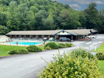 Outdoor pool and pool chairs surrounded by fencing, one story building with exterior guest room entrances, hotel entrance, parking spaces, shrubs and grassy areas