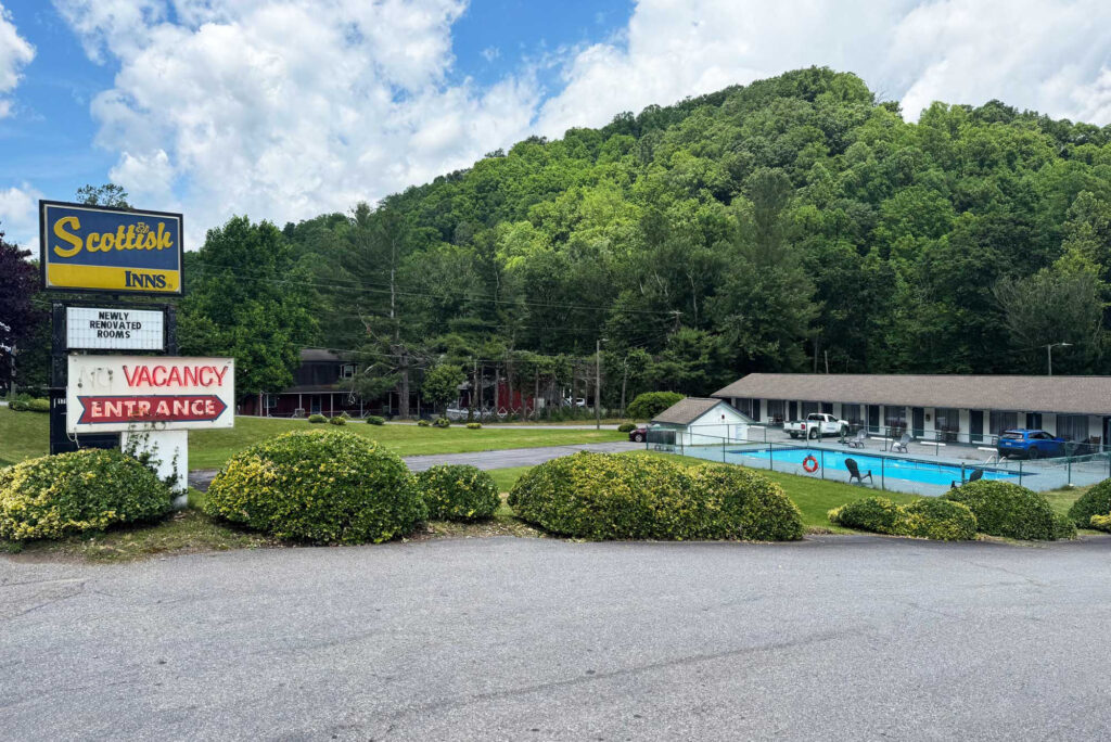 Hotel entrance, brand signage, landscaping with small bushes and grassy areas, fenced in outdoor pool, one story building with exterior guest room entrances, parking spaces