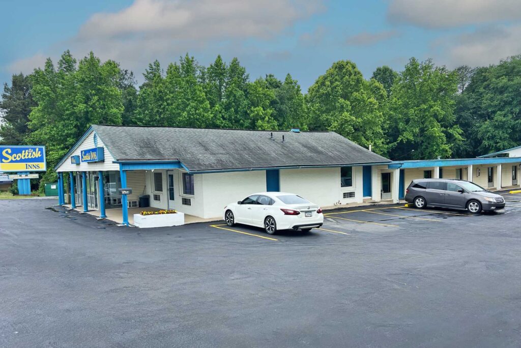 SNCMO-Exterior-100 Single story building with trees behind, hotel entrance with canopy and tub with flowers, exterior guest room entrances with covered walkway, parking spaces,