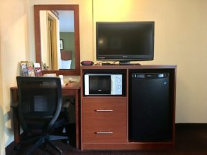 Desk with office chair, wooden unit with fridge, microwave, flat screen tv and clock, wall mounted mirror