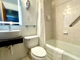 Vanity unit, illuminated mirror, hair dryer, small shelf, toilet, shower tub with bath mat, shower curtain and grab bars, tiled flooring