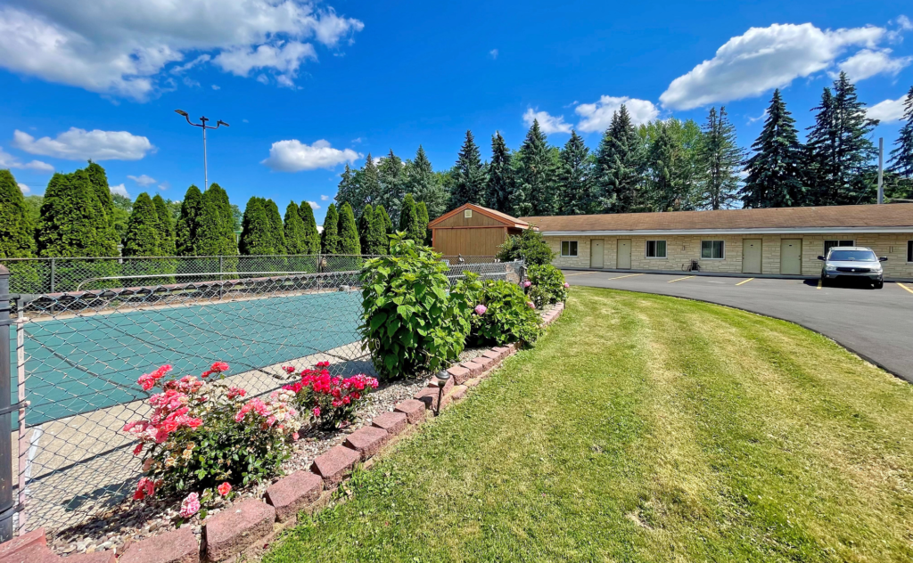 fenced in guest exterior pool with pool cover in place, flower shrubs, pine trees surround the pool, grassy area, one story building with exterior room entrances, parking spaces