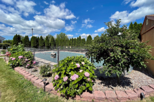 Fenced in guest exterior pool with pool cover in place, flowering shrubs, pine trees and grass around the pool fence