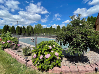 Fenced in guest exterior pool with pool cover in place, flowering shrubs, pine trees and grass around the pool fence