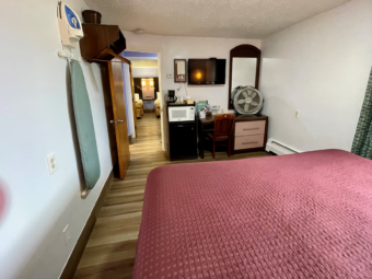 King bed, wall mounted ironing board and iron, hanging rail with hangers, fridge, microwave, small vanity table with drawers and chair, wall mounted flatscreen TV, mirror, fan, laminate flooring, doorway showing guest room with two queen beds acroos small hallway