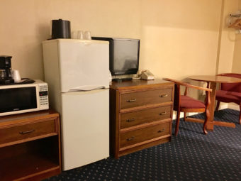 Wooden unit with storage space and drawer, microwave and coffee maker, Fridge with ice bucket and disposable cups, wooden unit with drawers, flat screen tv and telephone, small table and two upholstered chairs and carpet flooring