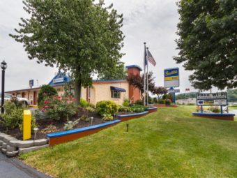 Hotel entrance, flower beds with flowering plants, bushes and trees, brand signage, parking spaces and grassy area