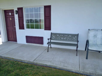 Exterior room entrance, concrete walkway, garden bench and chair, grassy area