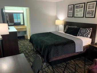King bed, art images, night stand with telephone and bedside lamp, easy chair, desk with chair, wooden unit with flatscreen TV, alcove with vanity unit, mirror with overhead light,carpet flooring