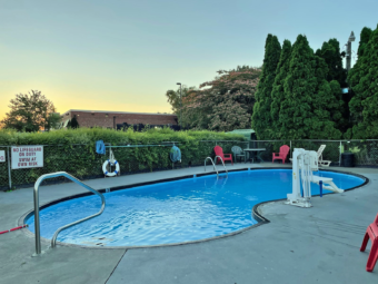 Outdoor pool with concrete poolside surrounded by fencing, privacy hedge and trees, ADA pool lift, patio chairs and table