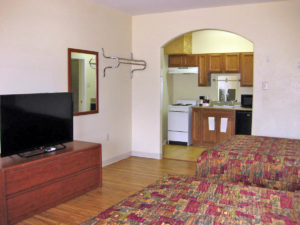 Two queen beds, wooden unit with flat screen tv, wall mounted mirror and hanging rail, laminate flooring, archway to kitchenette with stove, cabinetry, fridge and microwave