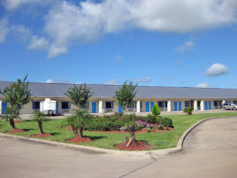 One story building with exterior room entrances, parking spaces, landscaped flower beds and grassy area with shrubs, flowers and small trees