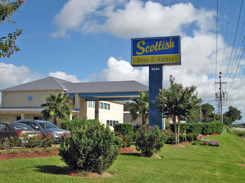 Hotel entrance with drive through canopy, brand signage, parking spaces, grassy area , landscaped flower beds and small bushes and trees
