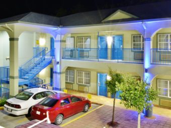 Two story building at night with exterior room entrances, walkways and stairs, aprking spaces, landscaped beds with small bushes and trees, parking spaces