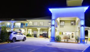 Hotel entrance displaying night time lighting with drive through canopy, small landscaping beds with bushes, parking spaces