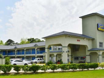 Hotel entrance with drive through canopy, two story building with exterior room entrances, walkways and stairs, parking spaces, grassy area with small bushes