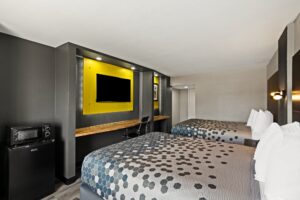 Fridge with microwave, wooden shelf below wall mounted TV, desk with office chair, art image, two queen beds, wall bedside lights, laminate flooring