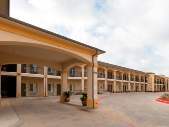 Two story building with exterior entrances to rooms, covered walkways and stairs, landscaping, potted plants, parking spaces