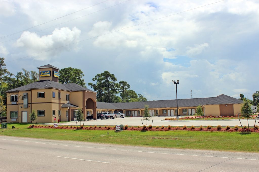 STXBN Exterior 108 Two story hotel entrance, one story attached building with exterior room entrances and covered walk way, parking spaces, landscaping with shrubs, grass and small trees