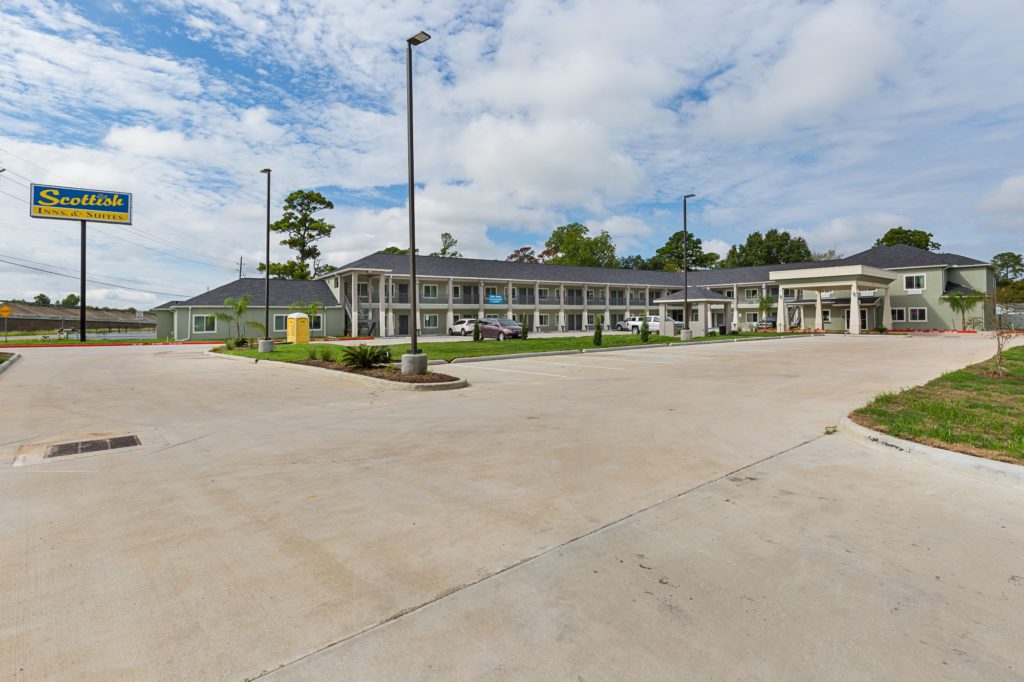 Hotel entrance with canopy drive through, brand signage, two story building with exterior room entrances, covered walkways and stairs, landscaping with grassy areas, shrubs and trees, parking spaces