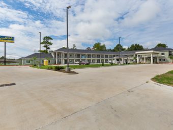 Hotel entrance with canopy drive through, brand signage, two story building with exterior room entrances, covered walkways and stairs, landscaping with grassy areas, shrubs and trees, parking spaces