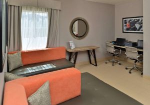 Two seating areas with cushions, wall mounted mirror, small occasional tablr, counter tops with computer monitors, keyboards and printer, wall mounted art, tiled flooring
