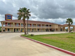 Two story building with exterior walkways to room entrances, hotel canopy entrance, parking spaces, grassy area and landscaping with small bushes