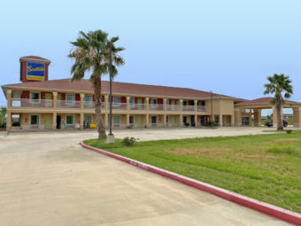 Two story building with exterior walkways to room entrances, hotel canopy entrance, parking spaces, grassy area and landscaping with small bushes