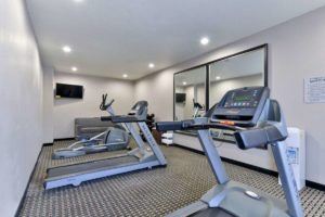 Fitness machines, wall mounted flat screen tv, sofa, weights, metal shelving with towels, large wall mounted mirrors, carpet flooring