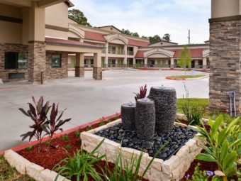 Hotel entrance with drive trhough canopy, two story building with exterior room entrances and covered walkways, lanscaping with shrubs, flowers and fountain, parking spaces