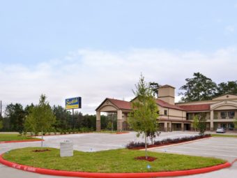 Hotel entrance with drive through canopy, brand signage, two story building with exterior room entrances and covered walkways, landscaping with shrubs, flowers, grassy area and parking spaces