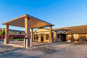 Drive through canopy, hotel entrance, external entrances to guest rooms, parking spaces, landscaping with grassy areas, white bench