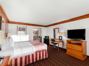 King bed, night stands with lamps and telephone, art images, fridge with microwave, desk with office chair and lamp, mirror, wooden drawer unit with TV, alcove with vanity unit and coffee maker, hanging rail with hangers carpet flooring