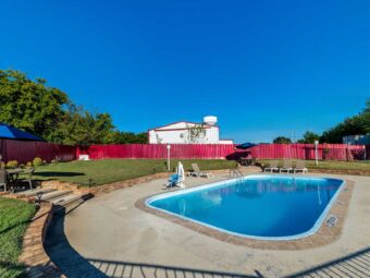 Outdoor pool with pool loungers, ADA pool chair, patio tables and chairs with umbrellas, concrete surround with steps to patio tables, grassy areas with lighting