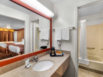 vanity unit with coffee machine and drink sachets, bathroom amenities, towel rail with towels, mirror with overhead light, doorway leading to showertub with bathmat and bathroom amenities, tiled flooring