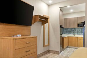 Wooden drawer unit, wall mounted TV, shelf with hanging rail, full length mirror, alcove leading to kitchenette area with full size fridge, sink hotplate, microwave and cabinets, laminate flooring