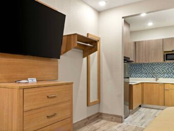 Wooden drawer unit, wall mounted TV, shelf with hanging rail, full length mirror, alcove leading to kitchenette area with full size fridge, sink hotplate, microwave and cabinets, laminate flooring