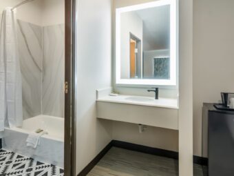 Alcove with fridge and shelf, vanity unit with backlit mirror, doorway to showertub with towels and shower curtain, tiled flooring