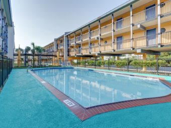 Two story building with covered parking overlooking a pool surrounded by fencing with concrete poolside and patio chairs and sun loungers