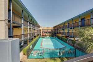 Two story building with covered parking overlooking a pool surrounded by fencing with concrete poolside and patio tables chairs and sun loungers