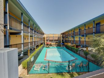 Two story building with covered parking overlooking a pool surrounded by fencing with concrete poolside and patio tables chairs and sun loungers