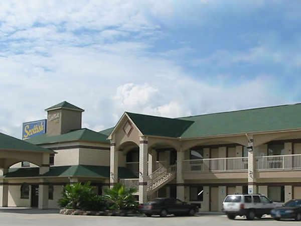STXHD Two story building with exterior room entrances, covered walkways and stairs, landscaping bed with shrubs and small trees, parking spaces