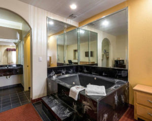 jacuzzi tub with towels and tiled surround, carpet flooring, alcove with vanity unit with wall mounted mirror with overhead light, wall mounted hair dryer, coffee maker, doorway to bathroom