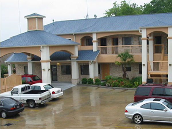 Hotel entrance with drive through canopy, two story building with exterior room entrances, covered walkways and stairs, landscaping with flowers, shrubs and palm tree, parking spaces