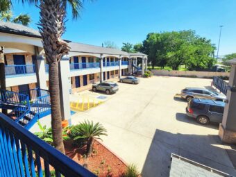 Two story building with exterior guest room entrances, covered walkways and stairs, parking spaces, landscaping with shrubs and trees