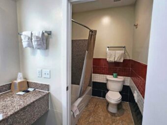 Vanity unit, back-lit mirror, towel rail with towels, doorway to bathroom with shower tub with shower curtain, toilet, towel rail with towels, tiled flooring